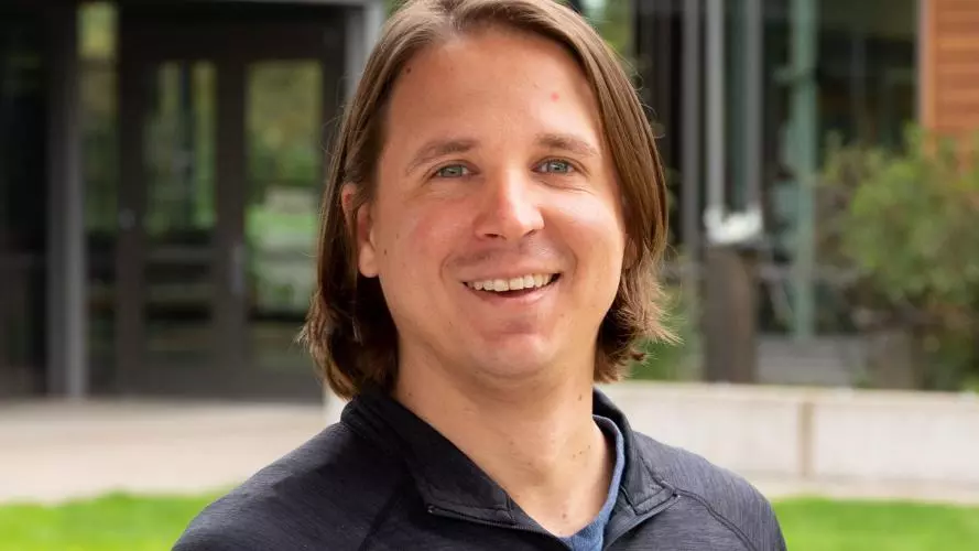 Sean has long straight brown hair and blue eyes. He is standing outside on some grass, wearing a dark grey lightweight sport jumper and smiling at the camera.