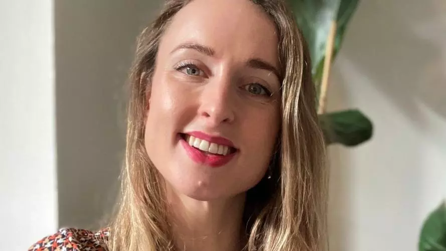 Adrienne has long blonde hair and is smiling at the camera. She's wearing a top with small white, orange and brown patterns.