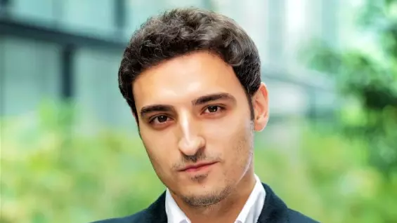 Alessio Cuozzo has short dark hair. He is wearing a white shirt and navy blazer, and is standing in front of a glass building.