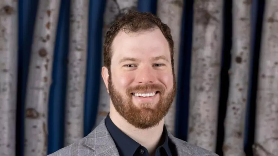 Alex Coccia smiles, standing in a grey suit jacket and dark-blue shirt 