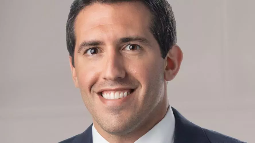 Alfonso is smiling at the camera. He has short brown hair and is wearing a navy suit with a white shirt and tie.