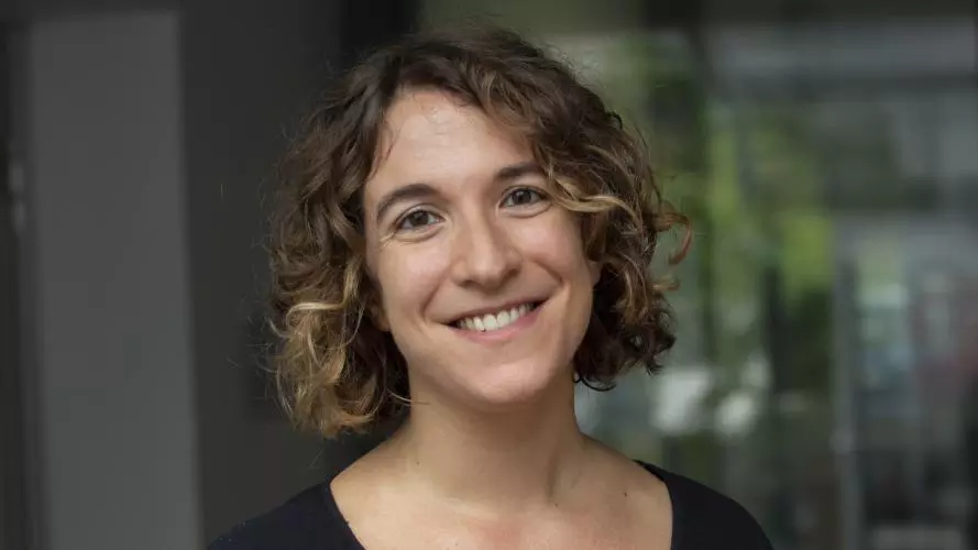 Arianna has short curly brunette hair. She is wearing a black t shirt and smiling at the camera