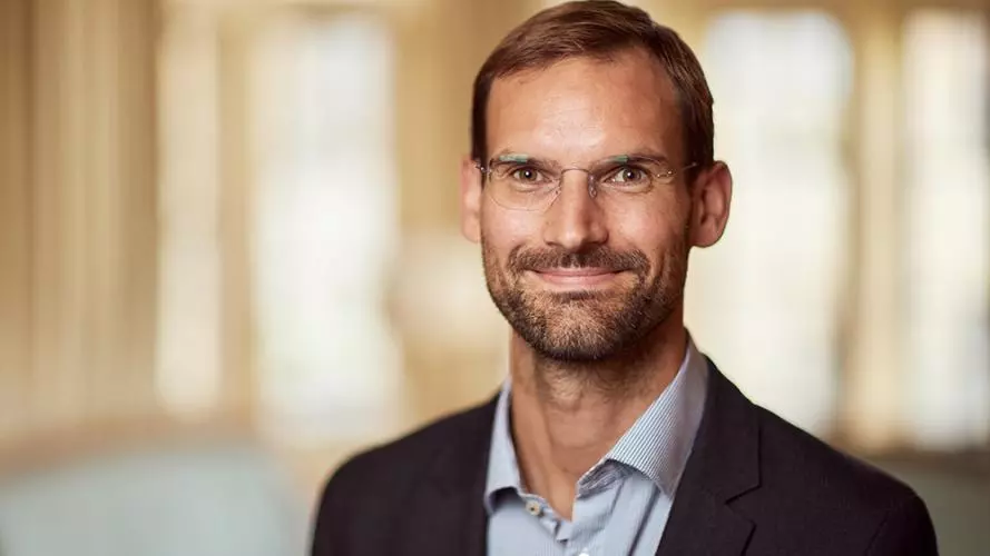 Man with broad smile wearing a light blue shirt and dark jacket. He has dark brown hair and beard.