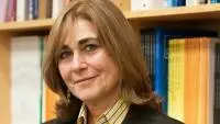 Jacqueline Bhaba is wearing a striped yellow shirt and blazer, standing in front of bookshelves
