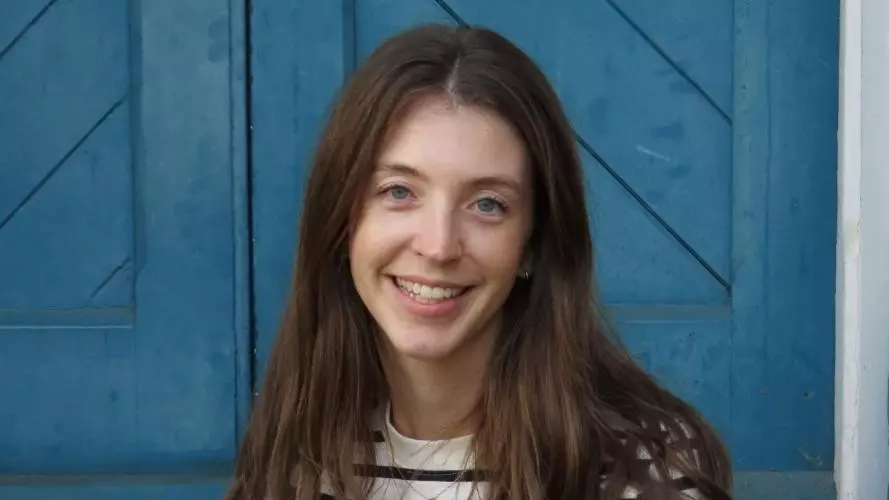 Bridget Steele has long brown hair and is wearing a black and white striped shirt. She is sitting on some outdoor steps in front of a blue door and smiling.