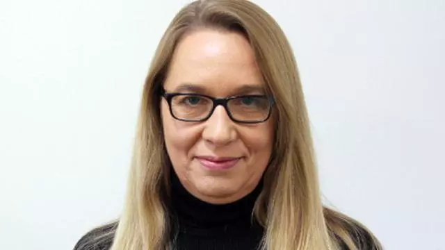 Woman with long blond hair, wearing dark rimmed glasses and black polo neck