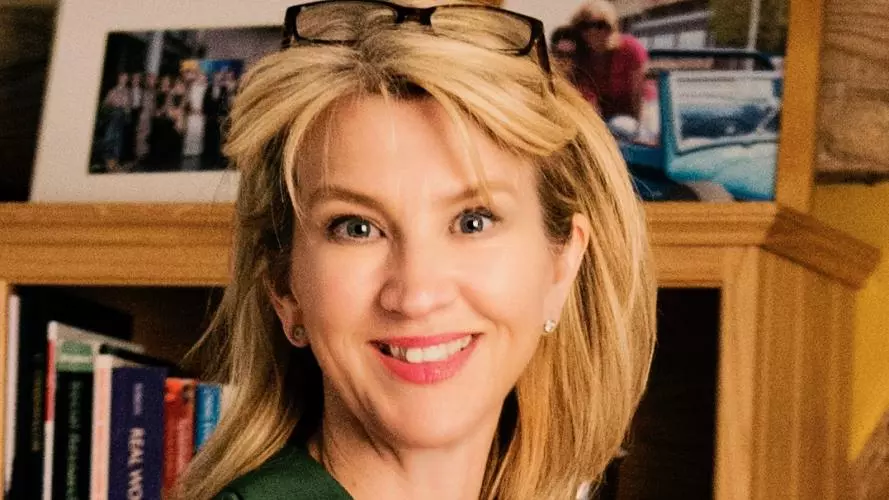 Caroline Greenhalgh wears a dark green shirt, has shoulder-length blonde hair and stands in front of a bookshelf. 