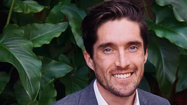 Man with dark hair who is smiling and wearing a white shirt and grey jacket. He is standing in front of a dark green hedge.