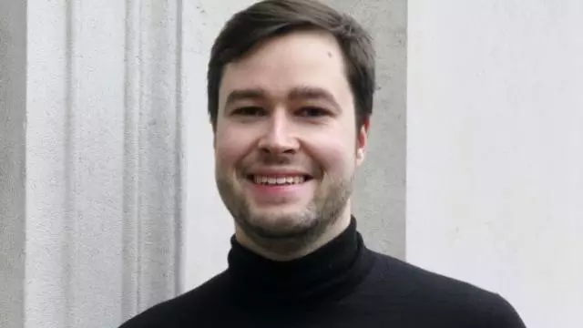 Christoph has short dark hair, a beard and is smiling at the camera. He's wearing a black polo neck top 