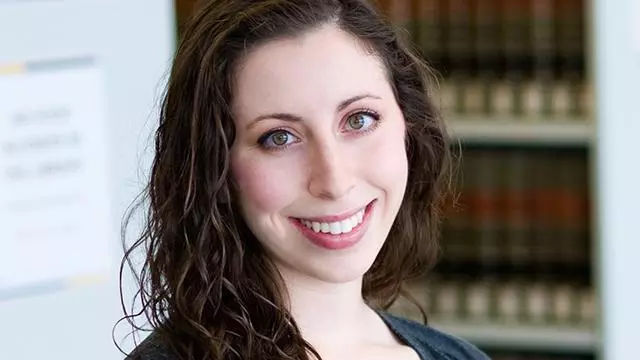 Alex has long dark curly hair. She is standing in front of some bookshelves, wearing a navy blue top and cardigan