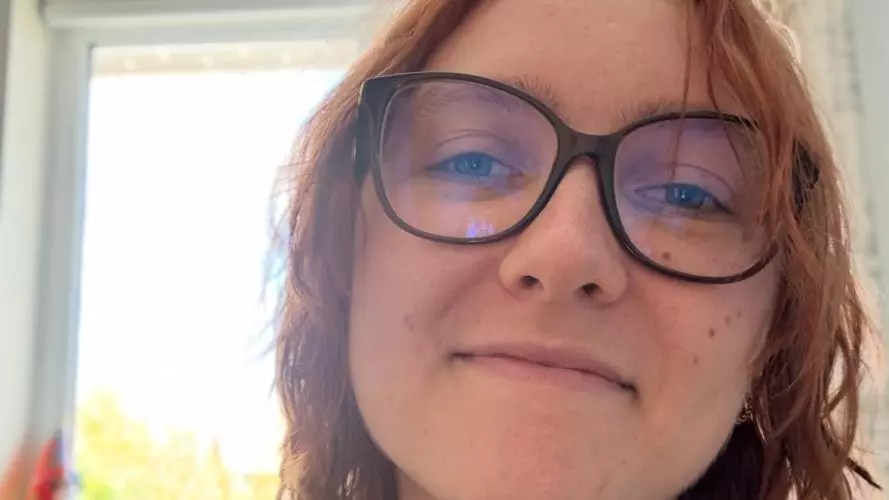 Ellie has shoulder length hair and is wearing brown glasses and a pale blue vest top
