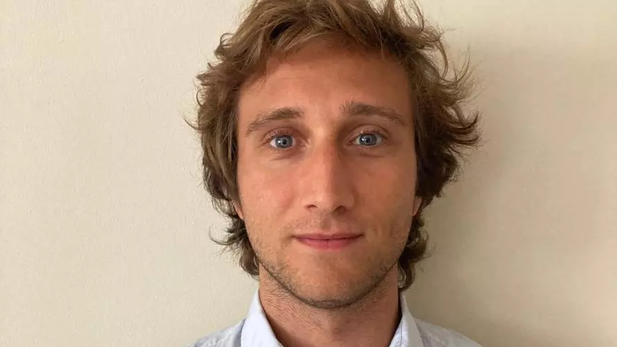 Enrico is standing in front of a wall looking at the camera. He has fair hair and is wearing a light shirt 