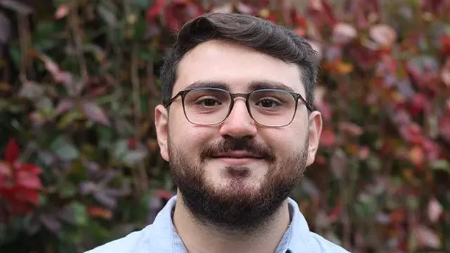 Ertugrul has short brown hair,  a brown beard and glasses. He's wearing a pale blue shirt