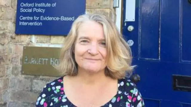 Smiling woman with light brown hair, wearing a patterned dress and standing in front of a dark blue door