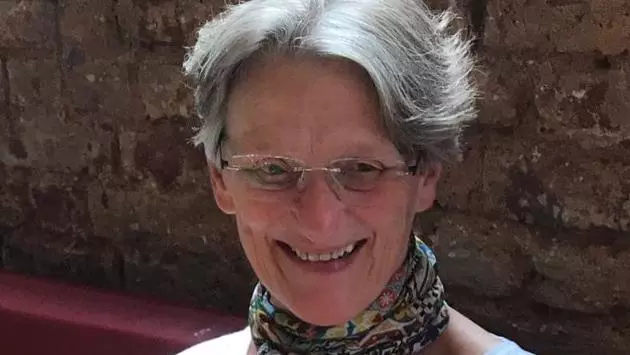 Professor Ward is looking slightly downwards and smiling. She has short grey hair and is wearing rimless glasses, a patterned scarf and a white t-shirt. 