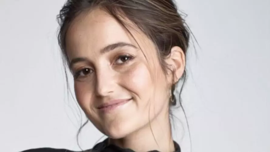 Eliana has brown hair in an updo and is wearing a high-neck draped black top. She is smiling at the camera.