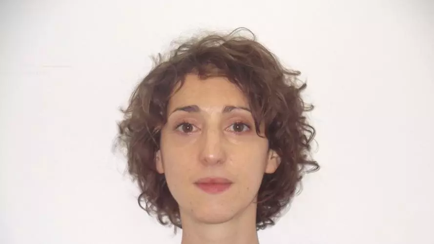 A headshot of Ilka Vari-Lavoisier. She has short curly brown hair. She is standing face-on to the camera, wearing a square-necked grey top with puffed sleeves.