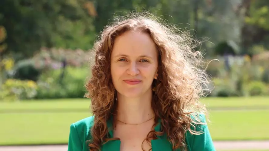 Jessica Lorimer has long curly brown hair. She is wearing a black dress and bright green blazer. She is standing outside in a large garden. 