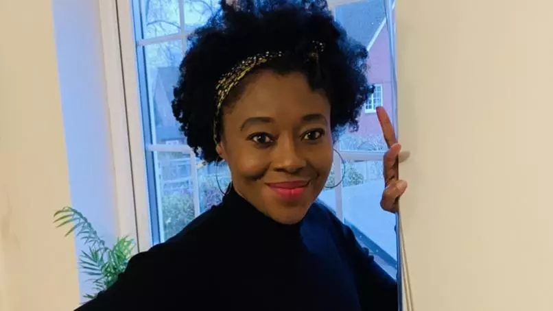 Isang Awah has black hair held back in a hair band, is wearing a black sweater and leading against a window