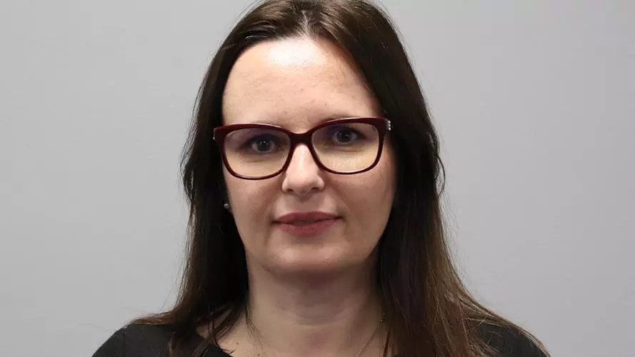 Woman with long brown hair, dark rimmed glasses and wearing a black top with necklace