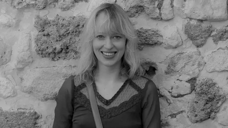 Janina has shoulder-length blonde hair and is smiling at the camera. She's wearing a dark-coloured top, and is standing against a stone wall