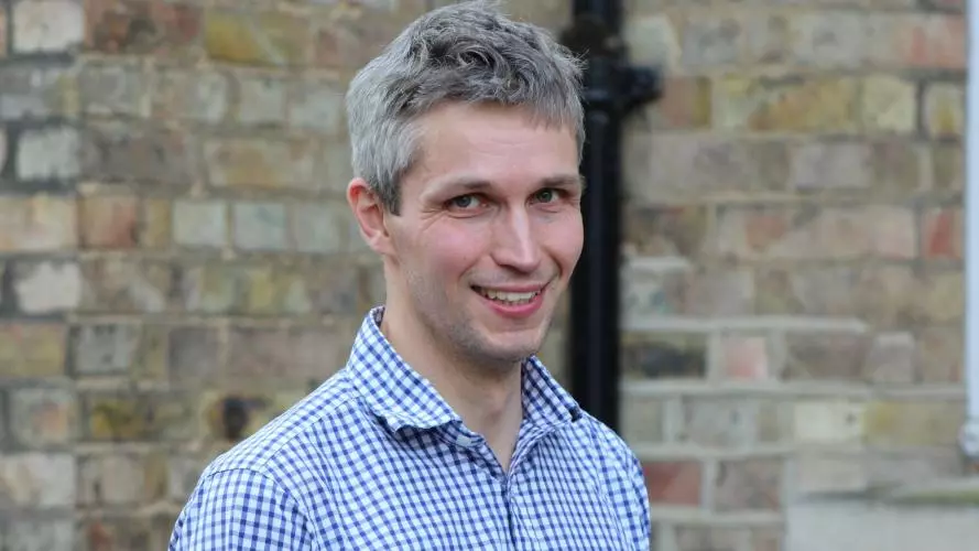 Jonathan is smiling at the camera. He has short grey hair and is wearing a blue and white checked shirt. 