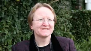 Judith has short fair hair. She is sat on a bench, wearing a dark purple outfit. 