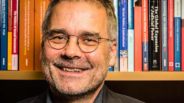 Martin Seeleib-Kaiser has short dark brown hair, glasses and is smiling at the camera. He's wearing a dark shirt and jacket and is sitting in front of a bookcase. 