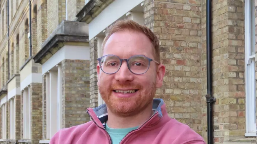 Dr Fanner has short fair hair and is wearing glasses. He is standing with his arms crossed, wearing a pink sweater. 