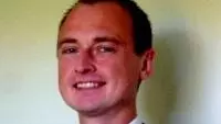 Pavelov has short brown hair. He is wearing a blue shirt with a white collar, dark tie and blazer