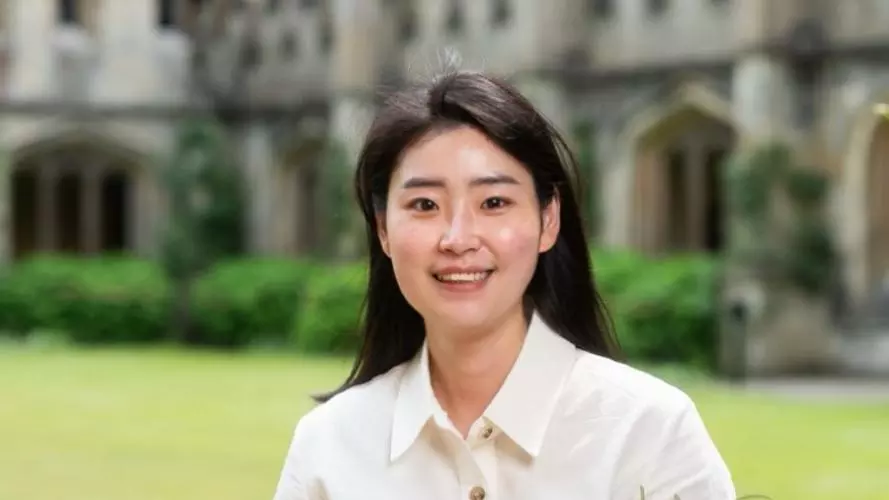 Seonwoo, with shoulder-length black hair, wearing a white shirt, stands on college grounds in front of a building, smiling at the camera.