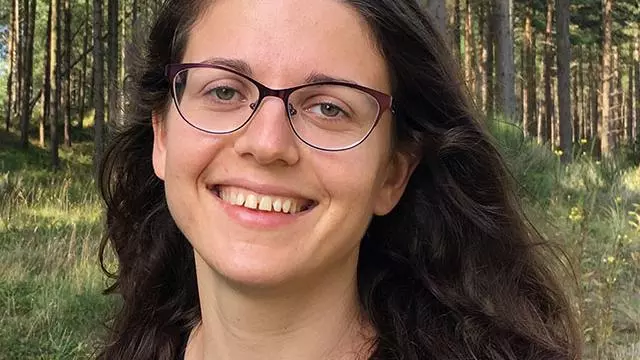 Yulia Shenderovich has long dark wavy hair, glasses and wearing a dark top. There is a background of trees behind her