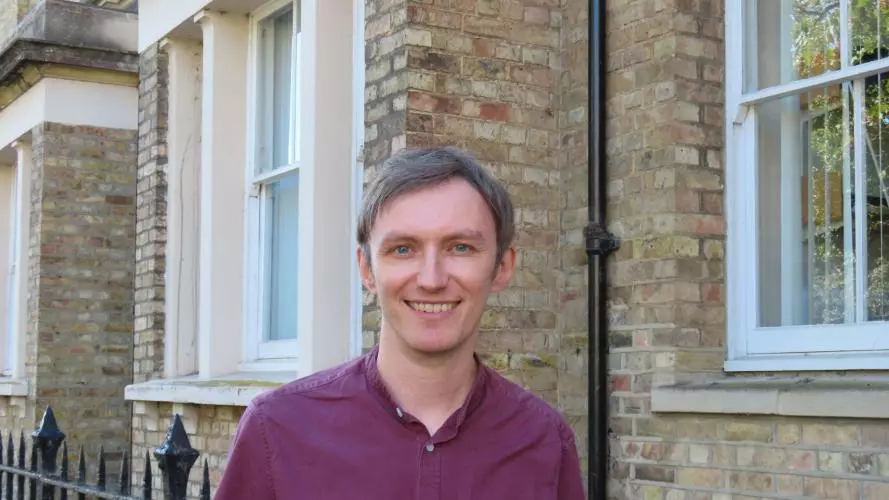 Robin has short fair hair and is wearing a short-sleeved burgundy shirt. He is standing outside in front of some light-brown brick buildings.