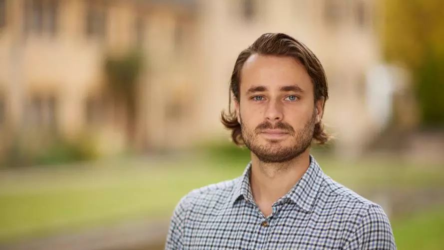 Roland has brown hair and beard, and is wearing a white checked shirt