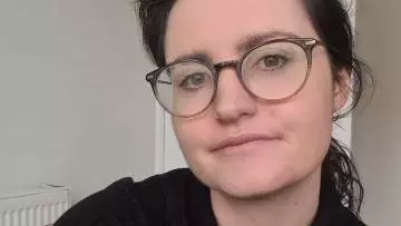Rosemary has long dark hair and brown eyes. She is wearing a black top and round glasses. Her hair is tied back into a ponytail. 