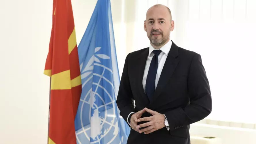 Mr. Benjamin Perks is wearing  a suit and tie standing in front of flags including at UN flag.