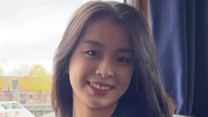 Yaxin has long dark hair. She is standing in front of a window and smiling. 