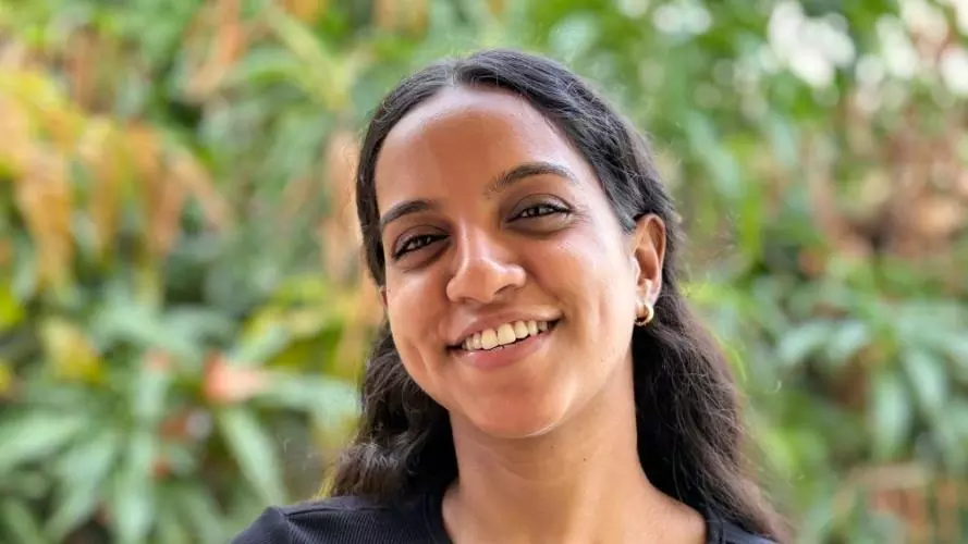 Tejashwari has long dark hair. She is wearing a black short sleeved t-shirt. She is standing in front of some foliage, smiling. 