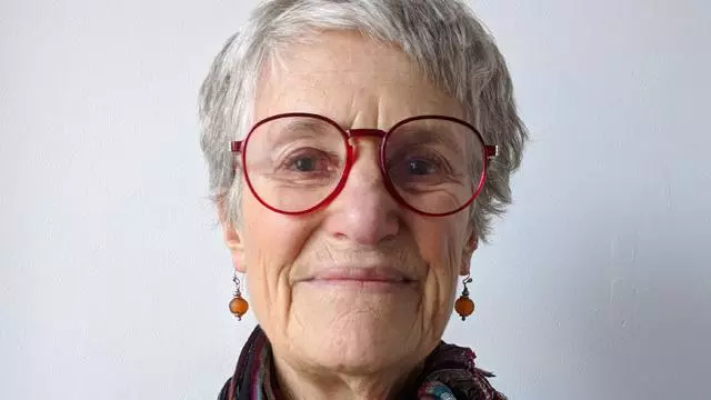 Woman with short grey hair, smiling and wearing red rimmed glasses and scarf around her neck.