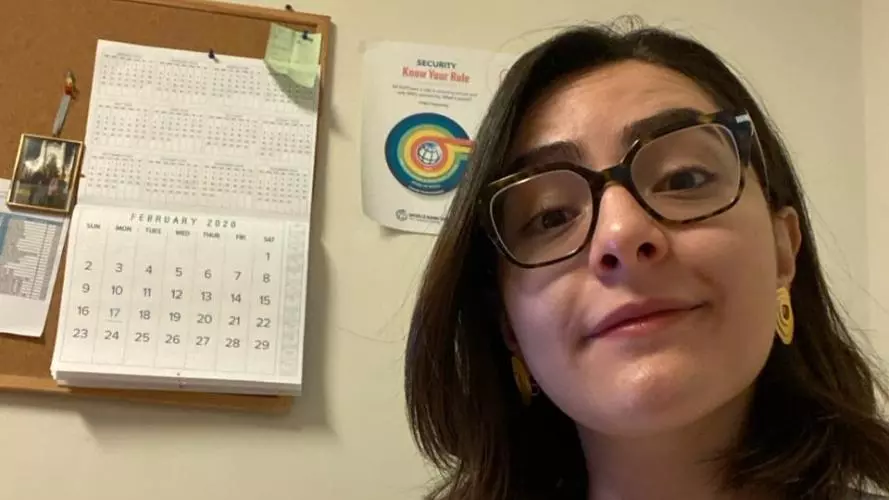 Nesma has shoulder-length dark hair, black-rimmed glasses and a white top. She is smiling at the camera