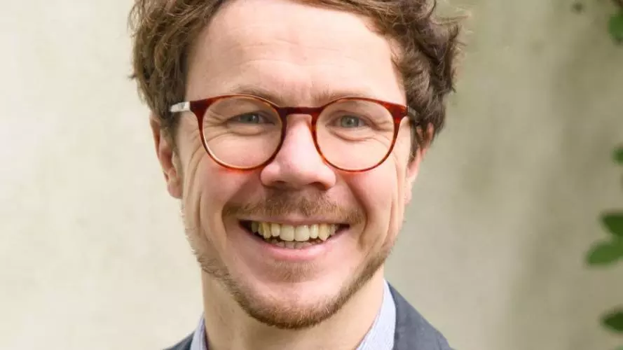 William Rudgard has short brown hair and round red glasses. He is wearing a shirt and blazer and smiling at the camera. 