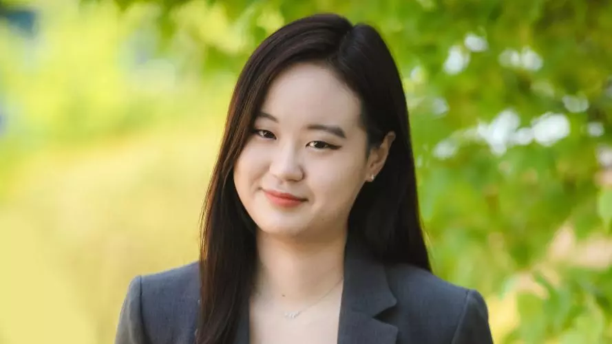 Ye Eun Ha has long dark hair and is wearing a black top and grey jacket. She's standing in front of a tree