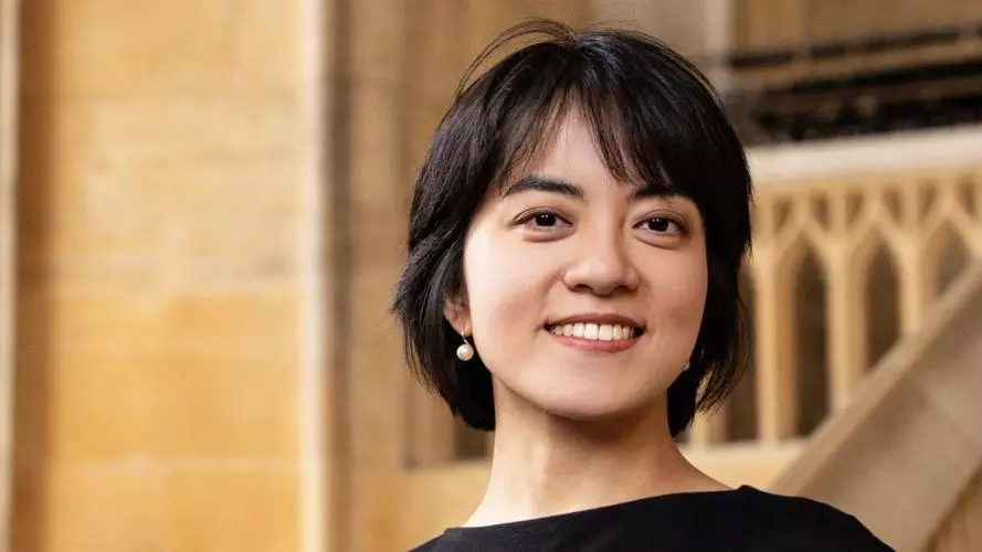 Yi Qie is wearing a black dress and pearl drop earrings with a short hair cut. She is smiling at the camera.