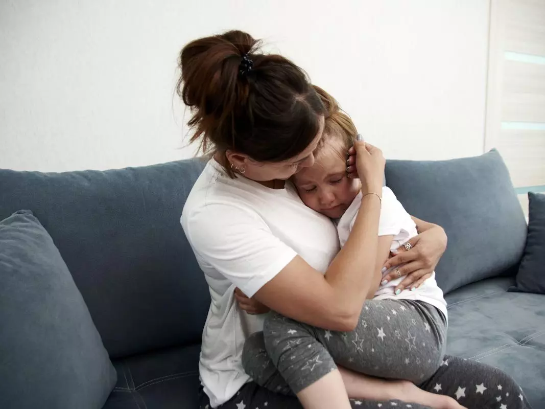 A mother sits on a grey sofa, hugging and comforting her young daughter