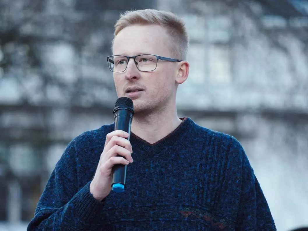 Benjamin is standing outside, wearing a navy jumper and speaking into a microphone.
