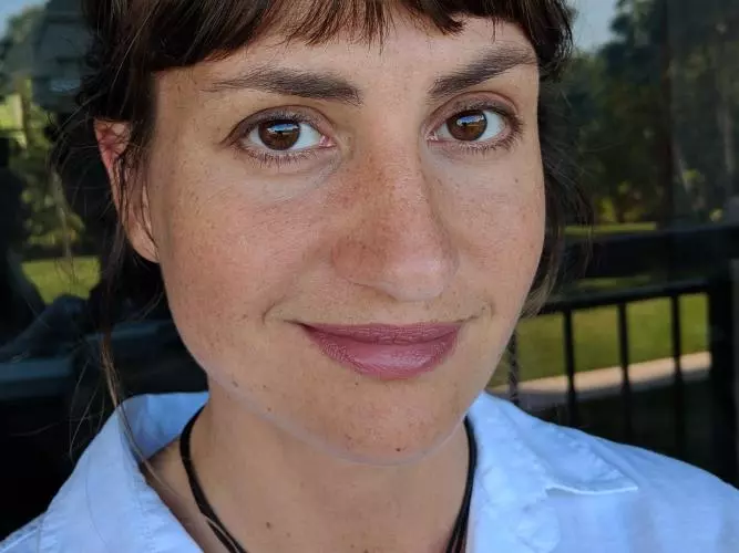 Smiling woman with short dark brown hair, wearing a light blue shirt and a black necklace. She is outdoors with trees and a railing in the background.