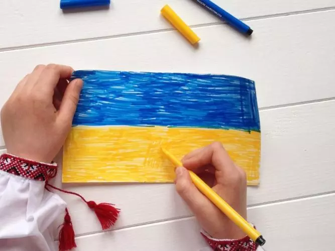 Hands colouring the Ukrainian flag blue and yellow on white paper, with coloured pencils nearby.