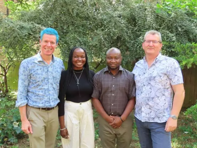 Four people smiling outdoors in front of greenery, standing side by side smiling at the camera.