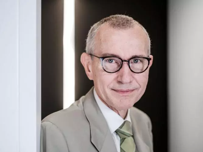 Professor Frank Vandenbroucke is wearing a light suit and round glasses. He is leaning against a light wall.