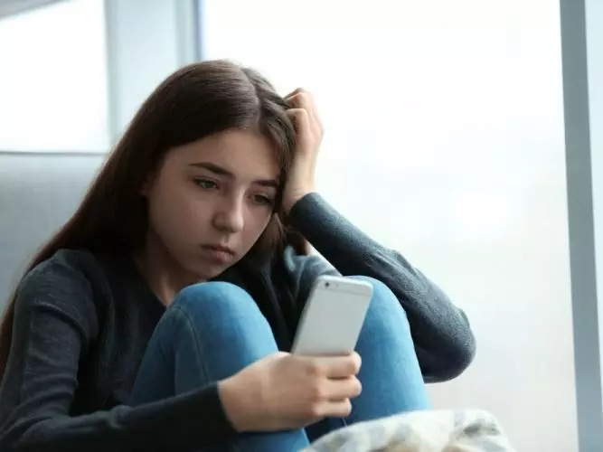 A young girl with long dark hair is sitting on a chair, looking at her phone with a sad expression. 